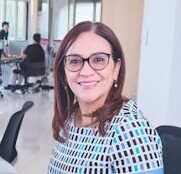 Confident woman smiling while working at an office desk with colleagues nearby.