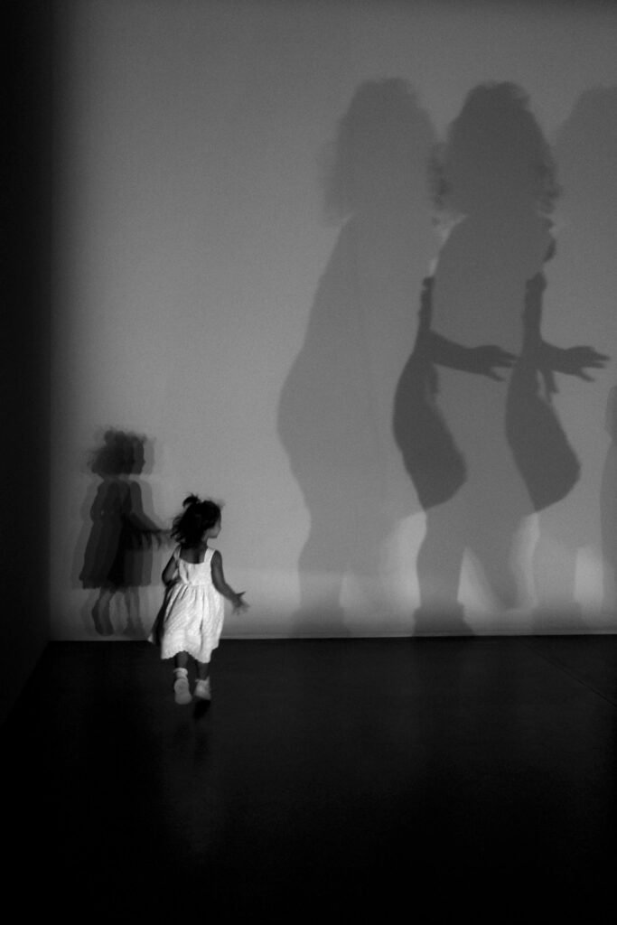 A child in a white dress runs towards projected shadows on a wall.